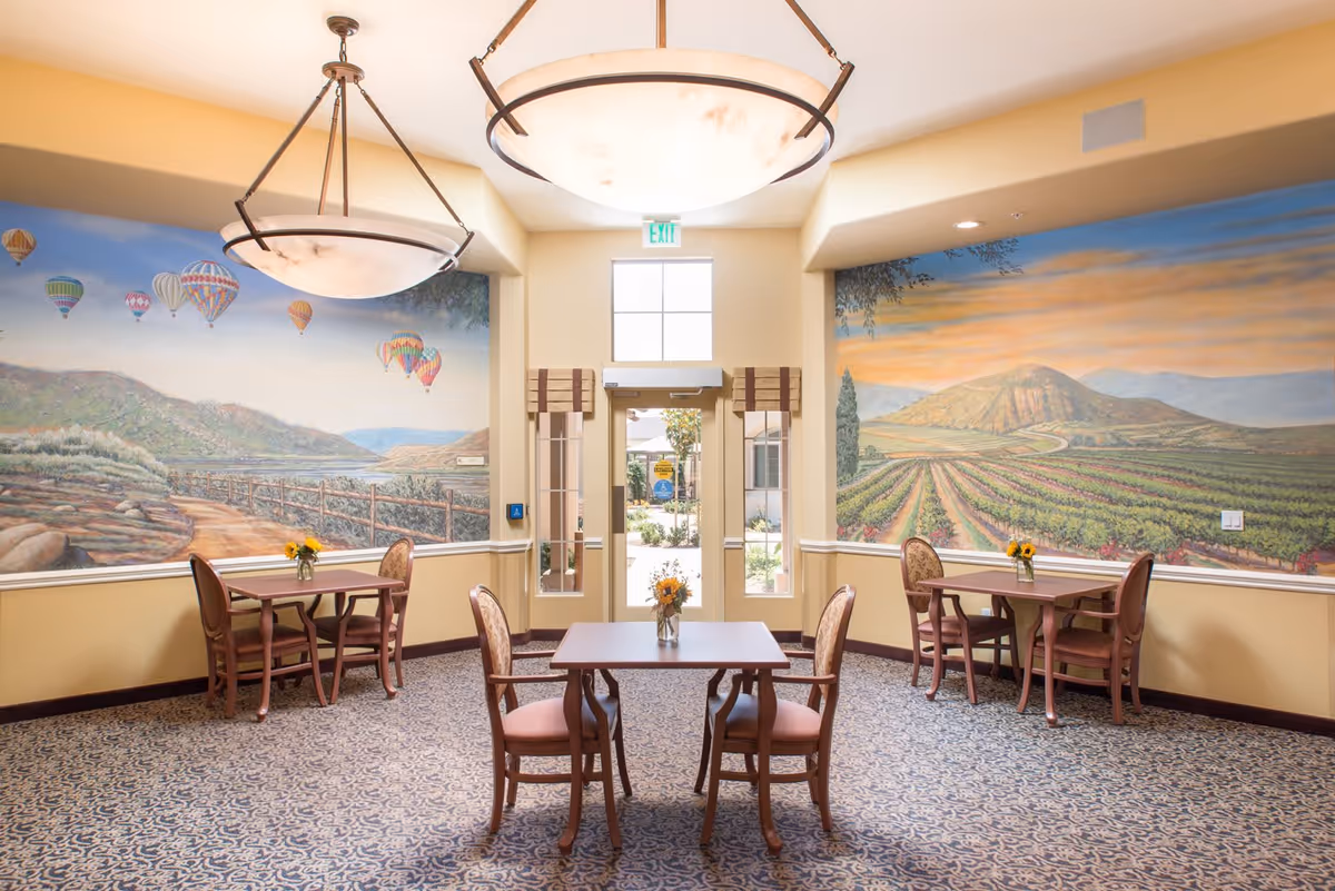 A bright dining area with three square wooden tables, each surrounded by four chairs with cushioned seats. Each table has a small vase with yellow flowers. The walls feature large murals depicting scenic landscapes with hot air balloons and vineyards. Two large ceiling lights hang above the tables, and a glass door with windows leads outside, allowing natural light to enter the room.