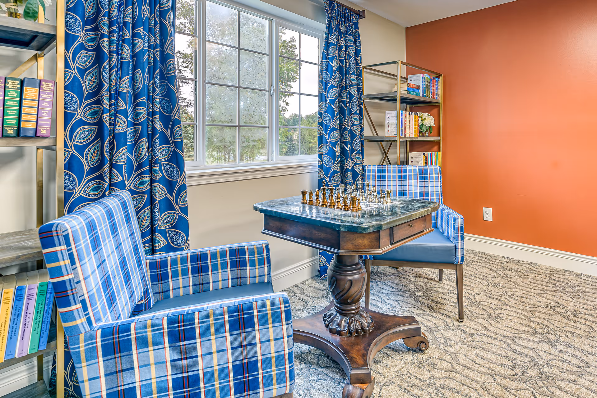 A cozy sitting area with two blue plaid armchairs facing a small wooden table with a chess set on it. The room has a large window with blue patterned curtains, a bookshelf filled with books, and an orange accent wall.