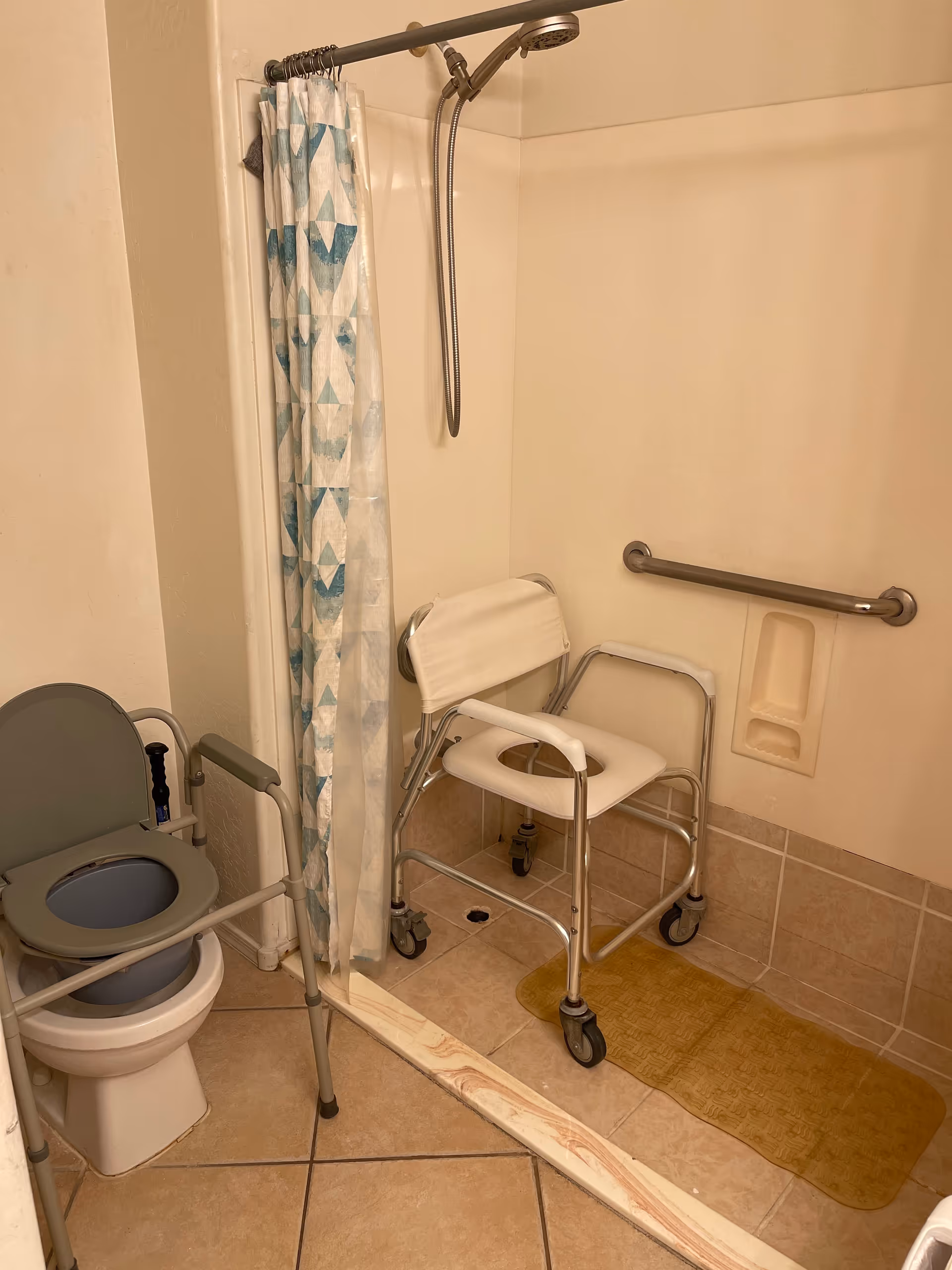 Bathroom with a toilet equipped with a raised seat and armrests, a shower area with a handheld showerhead, a shower curtain with a blue and white geometric pattern, a shower chair with wheels, a grab bar on the wall, and a yellow non-slip mat on the floor.