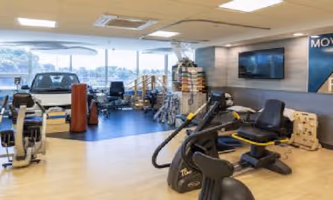 Interior view of a rehabilitation and nursing care facility room with various exercise and therapy equipment including stationary bikes, a treadmill, and other physical therapy tools. Large windows allow natural light to fill the space, and a flat-screen TV is mounted on the wall.