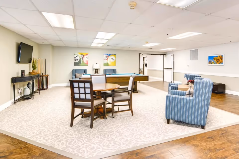 Bright communal recreation room with seating, a round table, a pool table, wall-mounted TV and patterned rug.