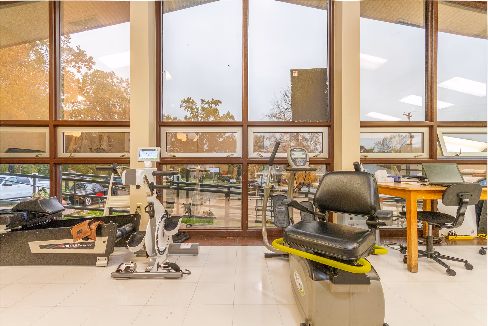 A bright room with large windows showing outdoor trees and parked cars. Inside, there are exercise machines including a recumbent bike and a rowing machine. A wooden table with office supplies and a black office chair is positioned near the windows.