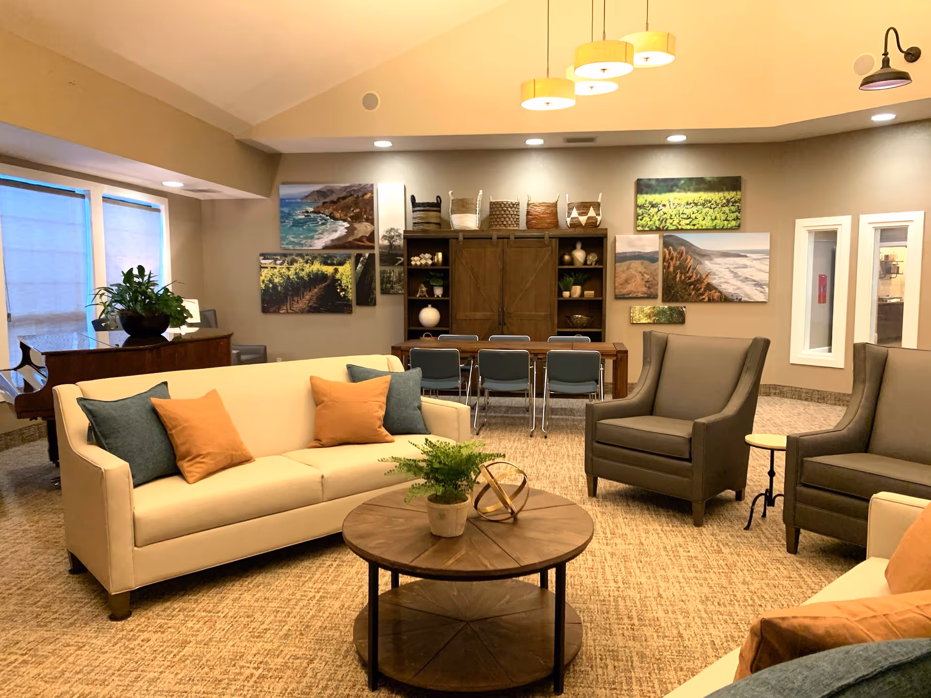 A cozy senior living common area with beige sofas adorned with orange and blue pillows, two gray armchairs, a round wooden coffee table with a small plant and decorative item, a wooden dining table with blue chairs, a piano near the window, and a wall decorated with landscape paintings and baskets on a wooden cabinet.