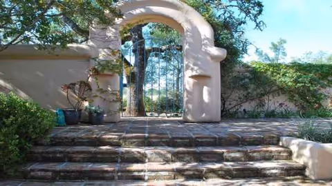 Stone steps lead to a rounded arch gate framed by greenery and stucco walls.
