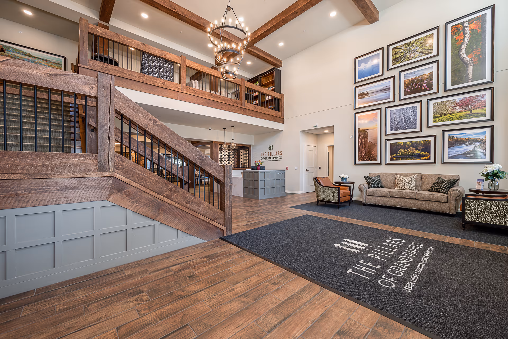 Spacious and well-lit interior lobby of The Pillars of Grand Rapids featuring wooden staircase with black railings, a reception desk, a seating area with a sofa and chair, and a wall decorated with multiple framed landscape photographs. The floor has a large mat with the facility's name and logo.