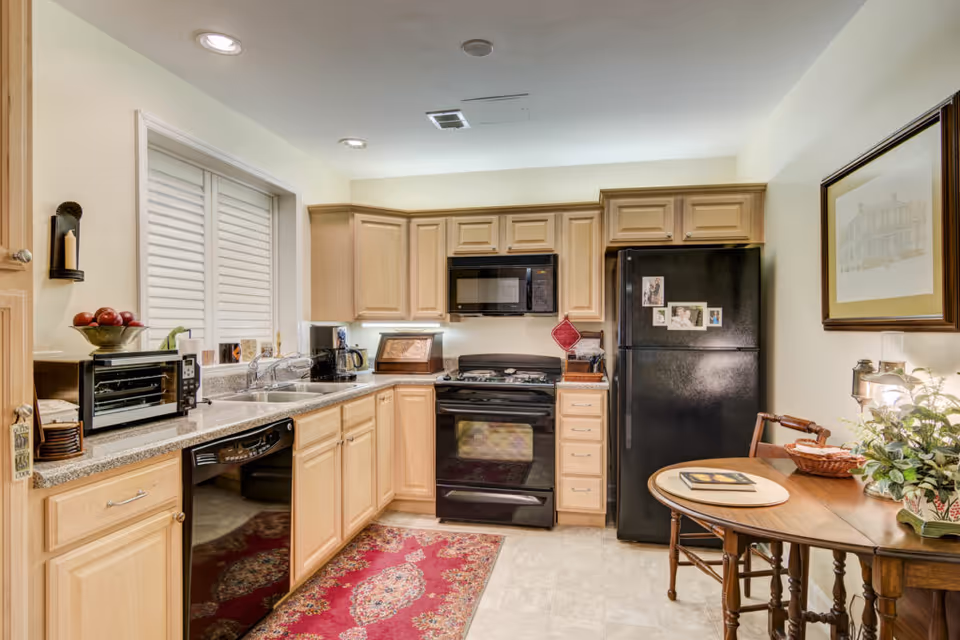 A bright kitchen with light wood cabinets, black stove and refrigerator, countertop appliances, and a small round dining table.