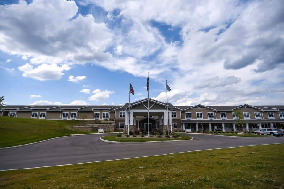 Front exterior view of Traditions of Beavercreek facility with a circular driveway, three flagpoles, and a partly cloudy sky.