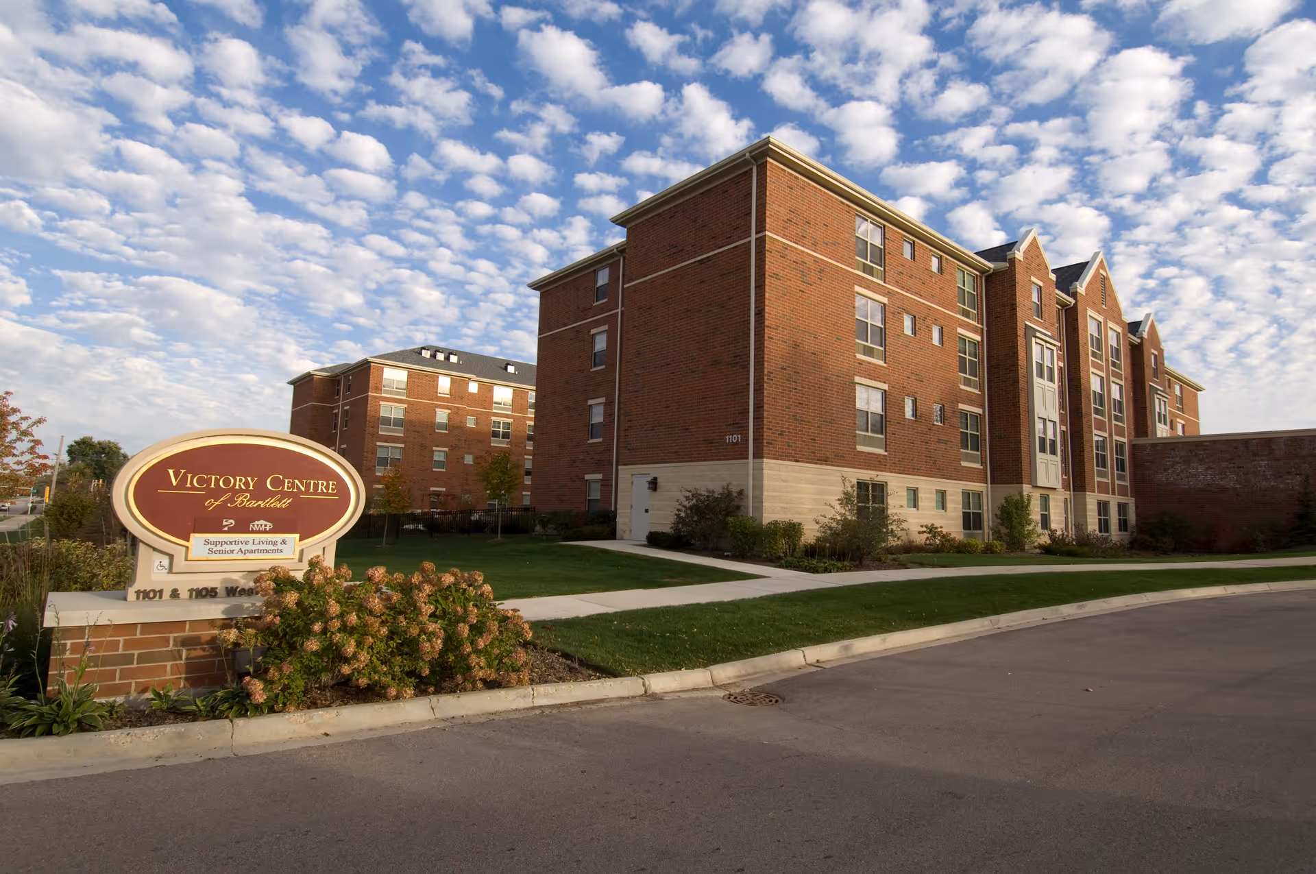 Exterior view of Victory Centre of Bartlett, showing two multi-story brick buildings with multiple windows, a well-maintained lawn, and a sign in the foreground that reads 'Victory Centre of Bartlett Supportive Living & Senior Apartments'. The sky is partly cloudy with scattered white clouds.