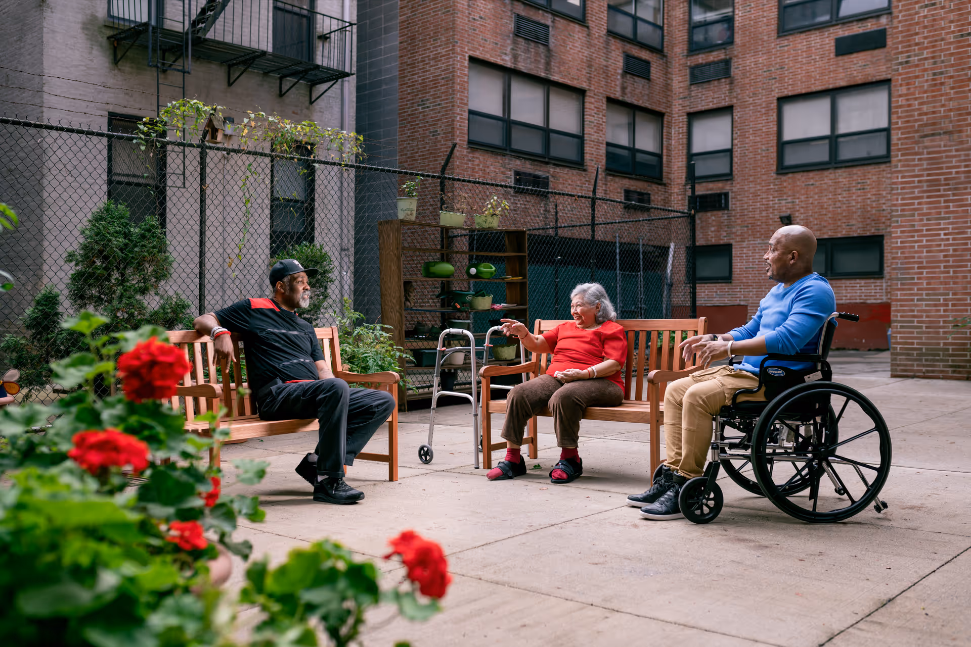 Three elderly individuals sitting and conversing in an outdoor courtyard area of a nursing facility. One person is seated on a wooden bench, another on a separate bench, and the third person is in a wheelchair. There are potted plants with red flowers in the foreground and a brick building with windows in the background.