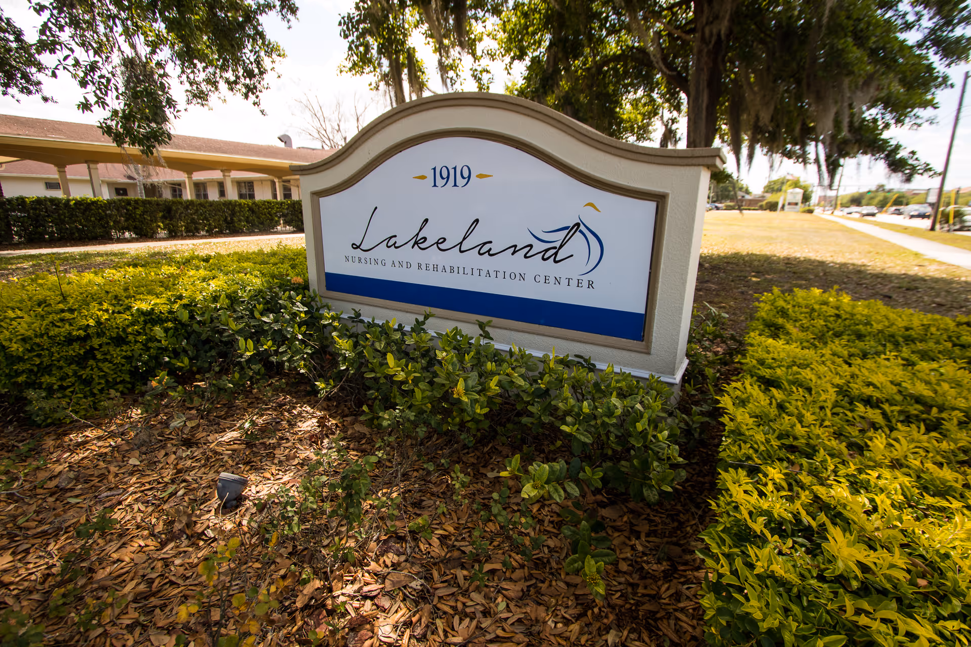 Outdoor view of a sign for Lakeland Nursing and Rehabilitation Center surrounded by green bushes and trees, with a building and sidewalk visible in the background.