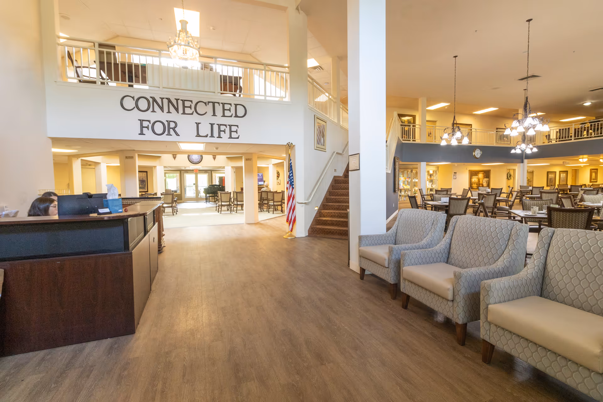 Spacious senior living facility interior with a reception desk, seating area, dining tables, and a large "CONNECTED FOR LIFE" sign on the upper wall.