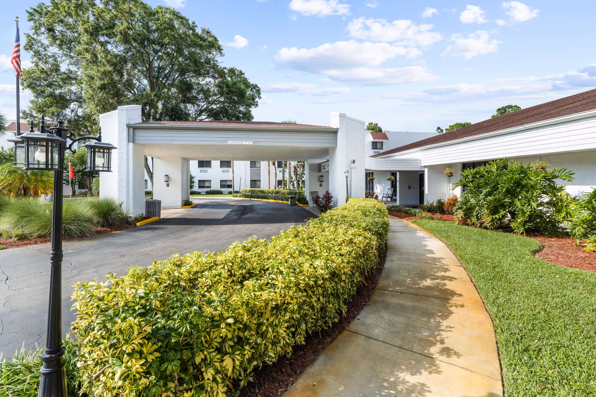 Exterior view of Ormond In The Pines Senior Living facility showing a driveway with a covered entrance, well-maintained landscaping including green bushes and grass, a sidewalk, a lamp post, and an American flag on a flagpole under a partly cloudy sky.