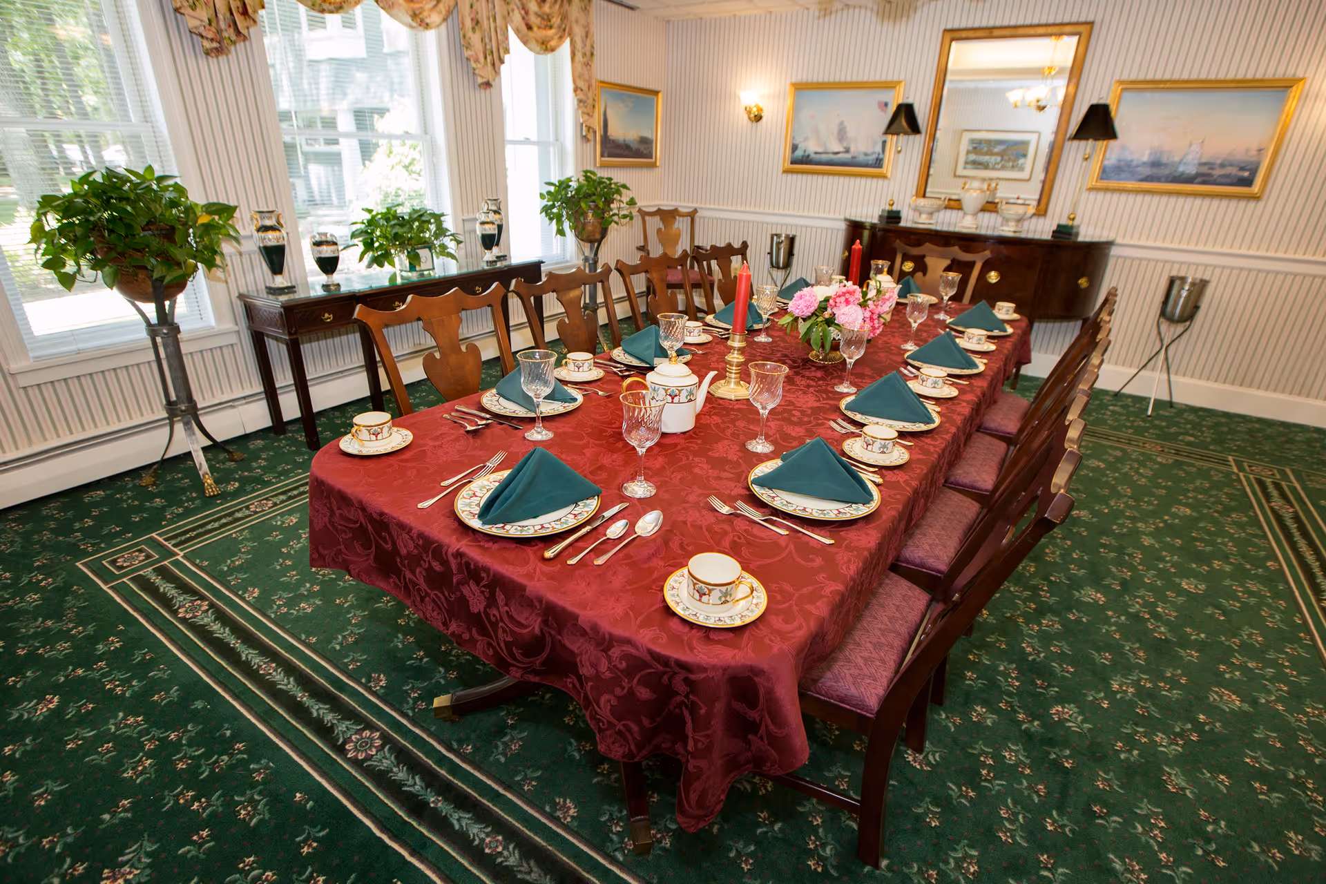 A formal dining room with a long wooden table covered by a red tablecloth, set with plates, cups, glasses, silverware, and green folded napkins. The room has green carpet with floral patterns, several wooden chairs with purple cushions, large windows with floral curtains, potted plants on stands and a side table, and framed paintings on the walls.