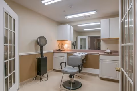 Interior view of a salon room with a salon chair in front of a counter with a sink, cabinets above and below the counter, a hair dryer on a stand, and fluorescent ceiling lights.