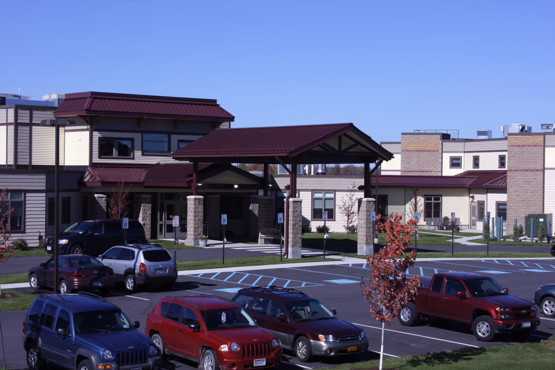 Front entrance and parking lot of a nursing home building with a covered porte-cochère and several parked cars.
