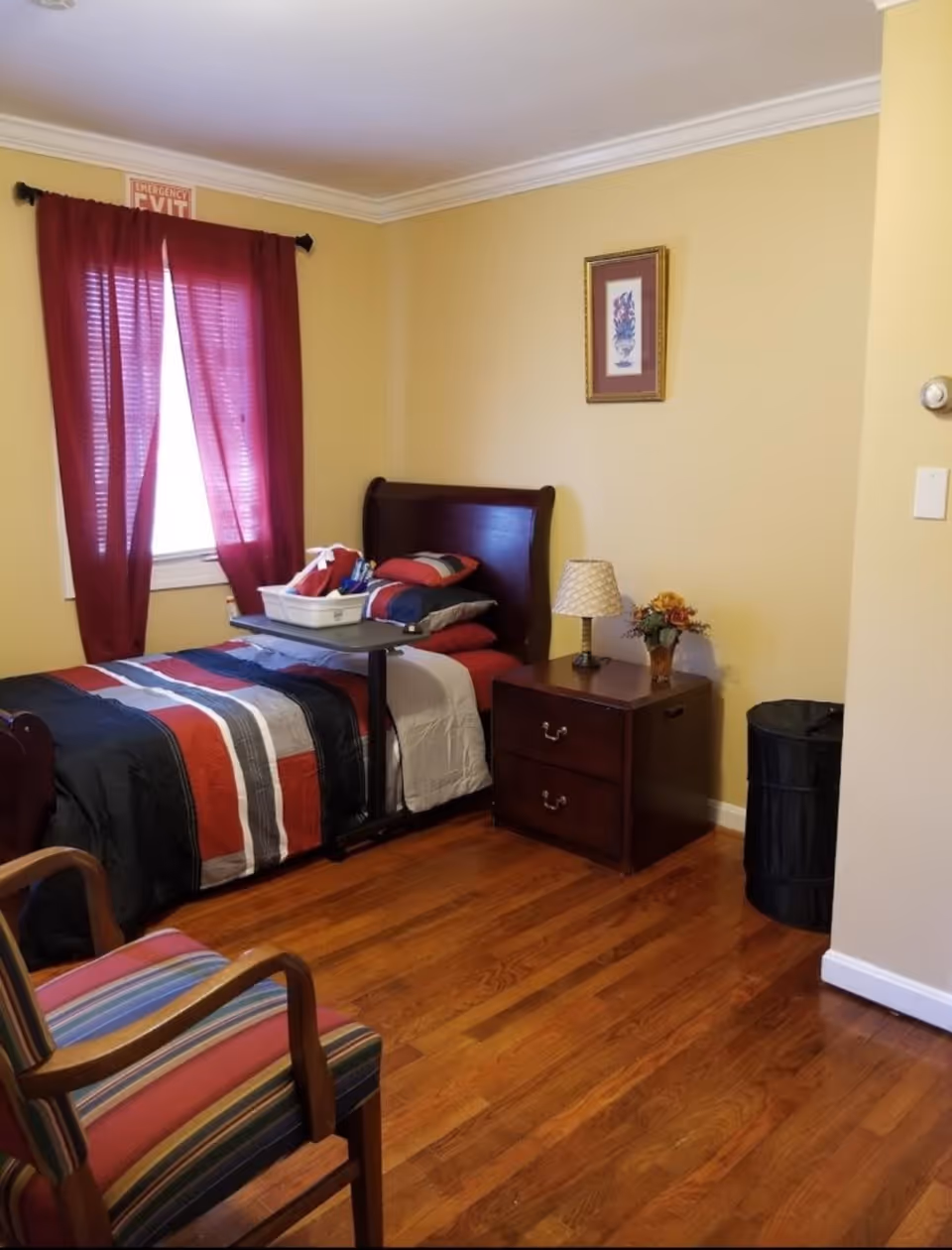 A cozy bedroom with a single bed featuring a dark wooden headboard and a striped bedspread in red, black, white, and gray. Next to the bed is a wooden nightstand with two drawers, a small lamp, and a vase with flowers. A window with red curtains is on the left wall, and a framed floral picture hangs on the yellow wall above the nightstand. A striped armchair is partially visible in the foreground, and a black laundry hamper is in the corner near the nightstand. The room has wooden flooring and an emergency exit sign above the window.