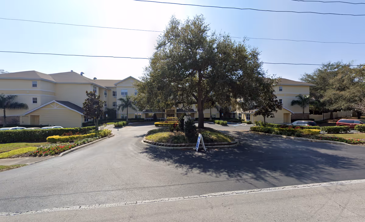Exterior view of The Colonnade at Beckett Lake senior living facility showing a three-story beige building with a central entrance, surrounded by landscaped greenery, palm trees, and a large tree in the middle of a circular driveway with parked cars on either side.