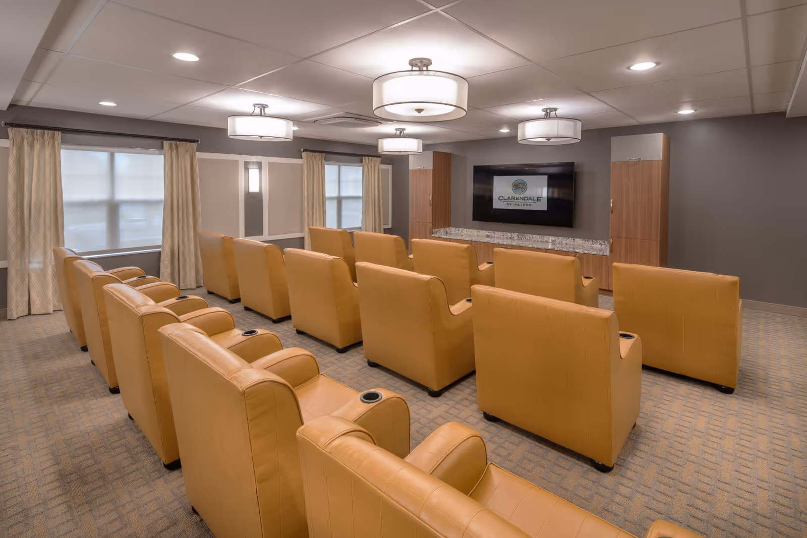 A cozy media room with three rows of tan leather armchairs facing a large flat-screen TV mounted on a gray wall. The room has carpeted flooring, two windows with beige curtains, and ceiling lights providing warm illumination. The TV screen displays the logo for Clarendale of St. Peters.