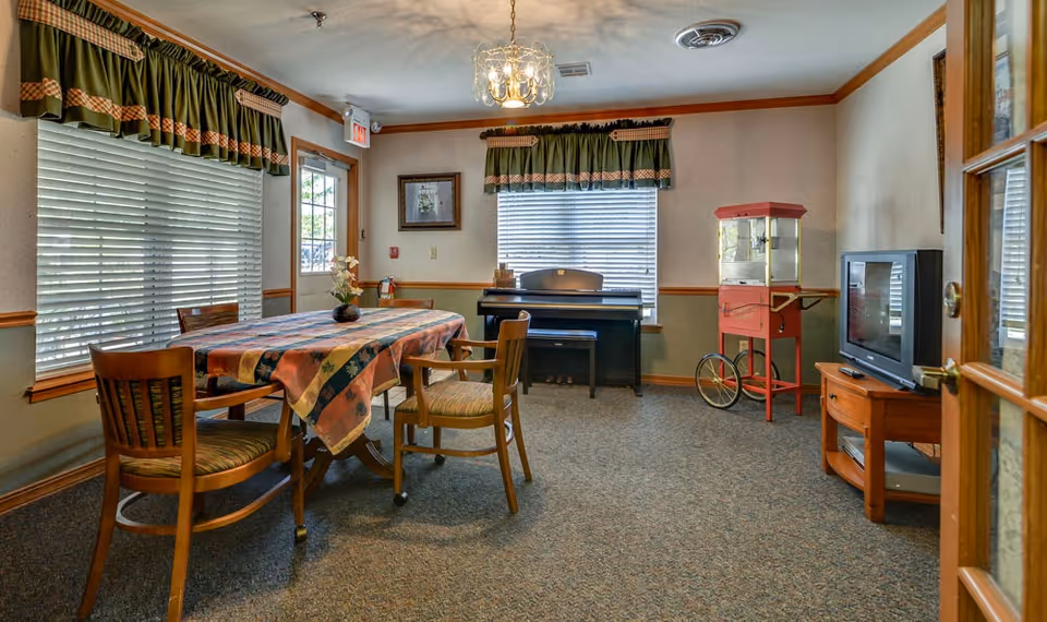 A cozy common room with a table covered by a patterned tablecloth surrounded by wooden chairs. The room has two windows with green valances, a piano against the far wall, a red popcorn machine on wheels, and a TV on a wooden stand. The walls are light-colored with wood trim, and there is a glass door on the right side.