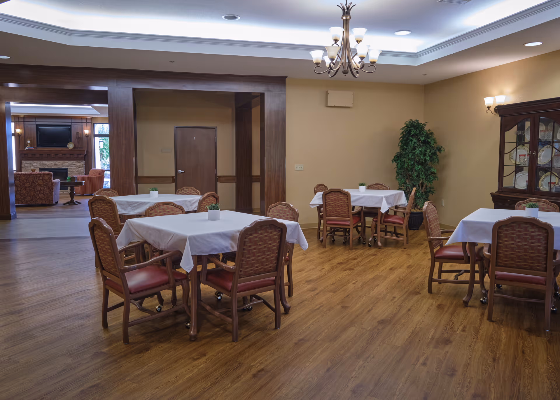 Dining room with several square tables covered in white tablecloths, wooden chairs, a china cabinet, potted plant, and a seating area visible through an open doorway.
