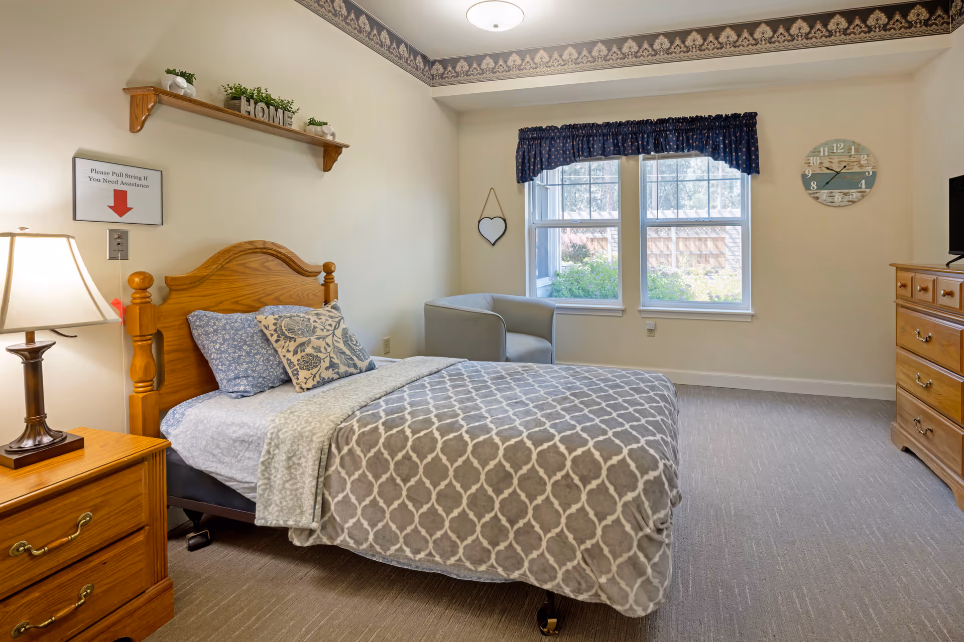 A cozy bedroom in a senior living facility with a wooden bed frame, patterned gray and white bedding, and two decorative pillows. There is a wooden nightstand with a lamp on the left side of the bed and a wooden dresser with a TV on the right. A gray armchair is placed near two large windows with blue valances, allowing natural light to brighten the room. A wall clock and a heart-shaped mirror hang on the walls, and a shelf above the bed holds small plants and a decorative 'HOME' sign. A sign on the wall instructs to pull a string for assistance.