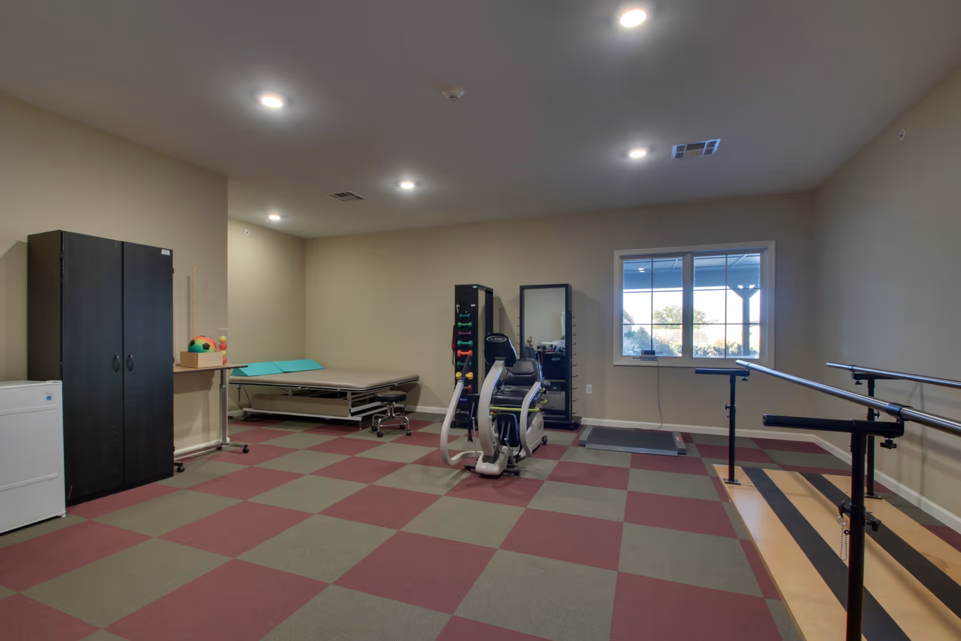 A spacious therapy or exercise room with checkered red and gray flooring, beige walls, and ceiling lights. The room contains a black cabinet, a white mini fridge, a therapy bed with a teal cushion, a rack with colorful dumbbells, a seated exercise machine, a full-length mirror, a treadmill, and parallel bars for walking support. A window on the right wall lets in natural light.