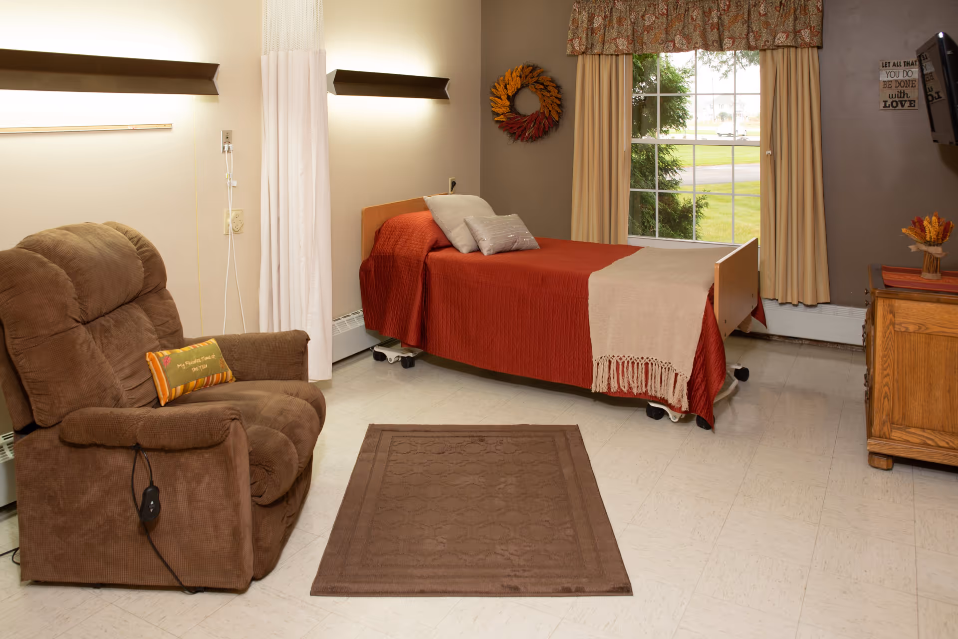 A cozy senior living room featuring a brown recliner chair with a small decorative pillow, a single bed with a red bedspread and beige throw blanket, a window with beige curtains and a floral valance, a wooden dresser with a small vase of flowers, and a wall decoration with a wreath and a framed quote.