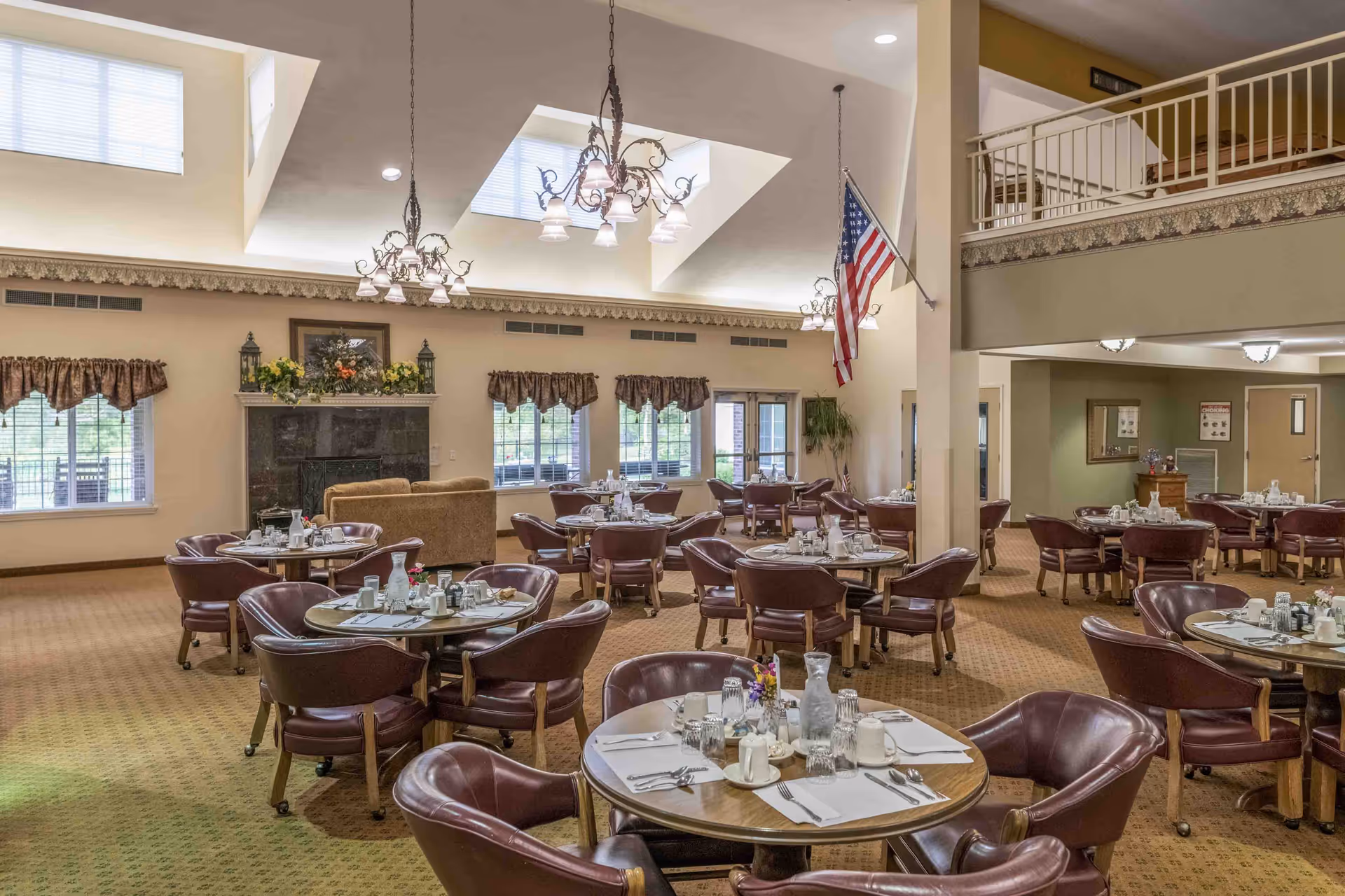 A spacious dining room with multiple round tables set with white placemats, silverware, glasses, and pitchers. The room features brown leather chairs, a fireplace decorated with flowers, large windows with valances, chandeliers hanging from a high ceiling with skylights, and an American flag mounted on a pillar.