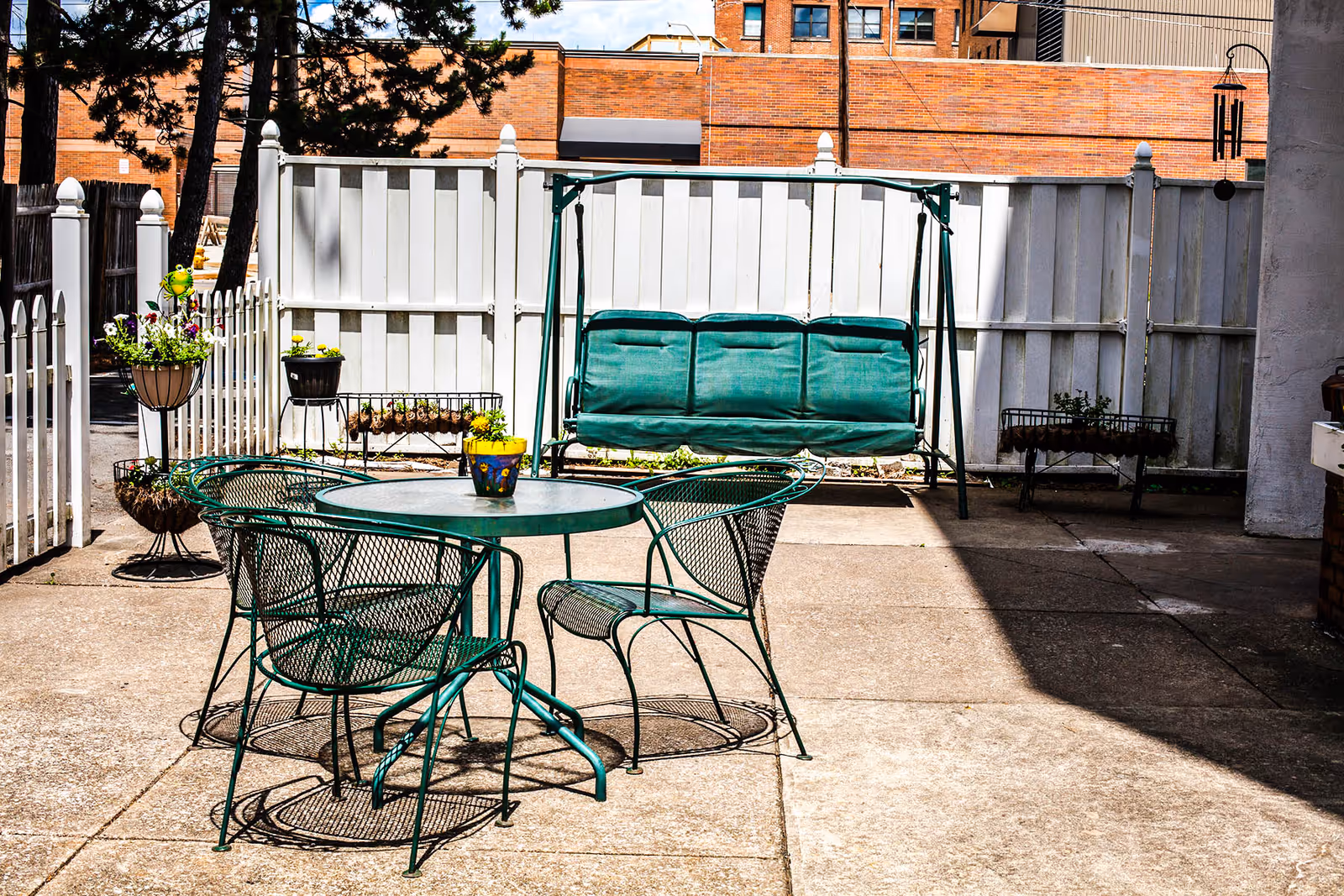 Outdoor patio with a green metal table and chairs, a green cushioned swing bench, and potted plants in front of a white fence.
