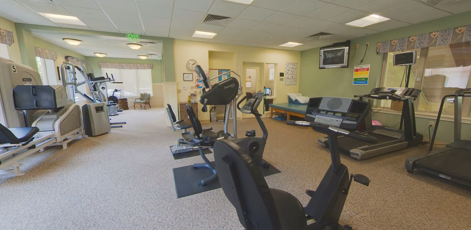Interior view of a fitness room in Ware Presbyterian Village featuring various exercise equipment including stationary bikes, treadmills, and weight machines. The room has carpeted floors, light green walls, windows with patterned valances, a wall-mounted TV, and a clock on the wall.