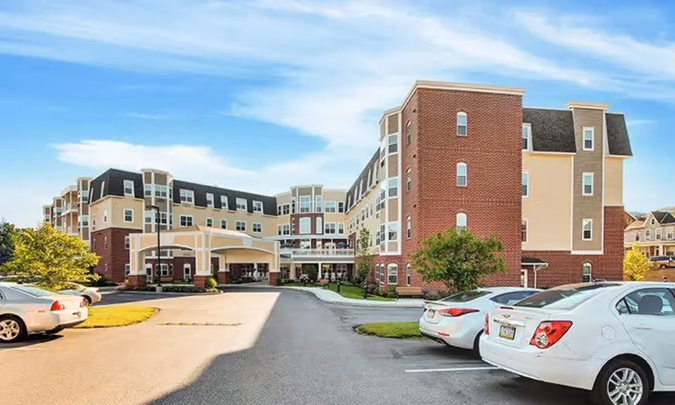 Front exterior of a multi-story brick-and-beige senior living building with a covered entrance and parked cars in the lot.