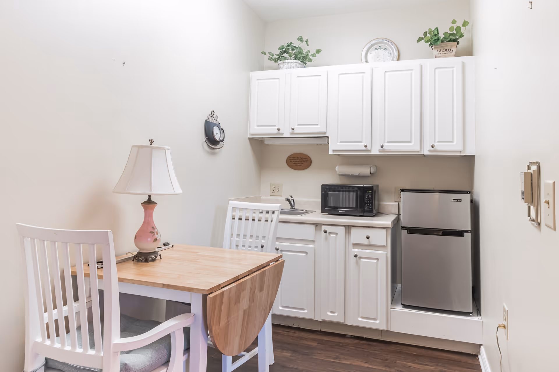 Small kitchenette area with white cabinets, a countertop holding a microwave and a small refrigerator. A wooden table with a pink lamp and two white chairs is positioned in front of the kitchenette. There are decorative plants and a plate on top of the cabinets.