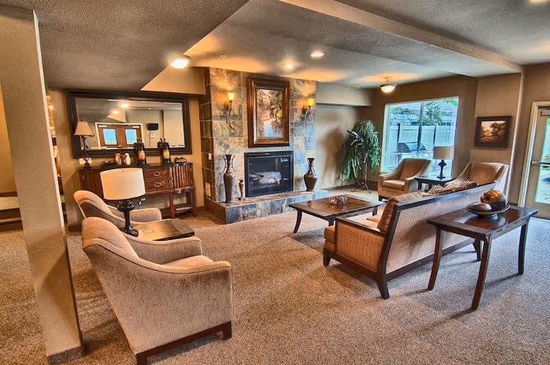 A cozy senior living facility common area with beige carpet and neutral-toned walls. The room features a stone fireplace with a framed painting above it, surrounded by two wall sconces. There are several comfortable chairs and a sofa arranged around a wooden coffee table. A large mirror hangs on the wall above a wooden sideboard with decorative items. A window and glass door allow natural light to enter the room, and a potted plant adds greenery to the space.