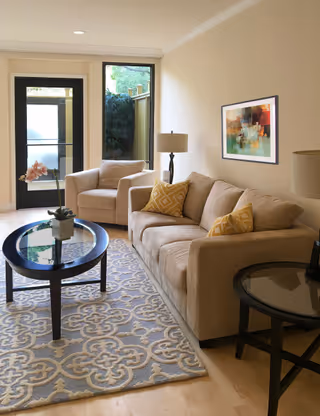 Bright living room with a beige sofa and armchair, patterned rug, round coffee and side tables, table lamps, and framed artwork near a glass door.