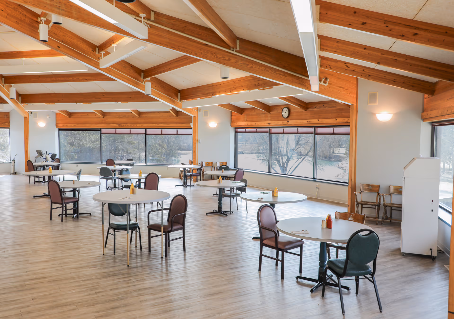 A spacious dining room with round tables and chairs arranged throughout. The room features large windows with views of trees outside, wooden beams on the ceiling, and light wood flooring. Each table has condiments like ketchup and mustard.