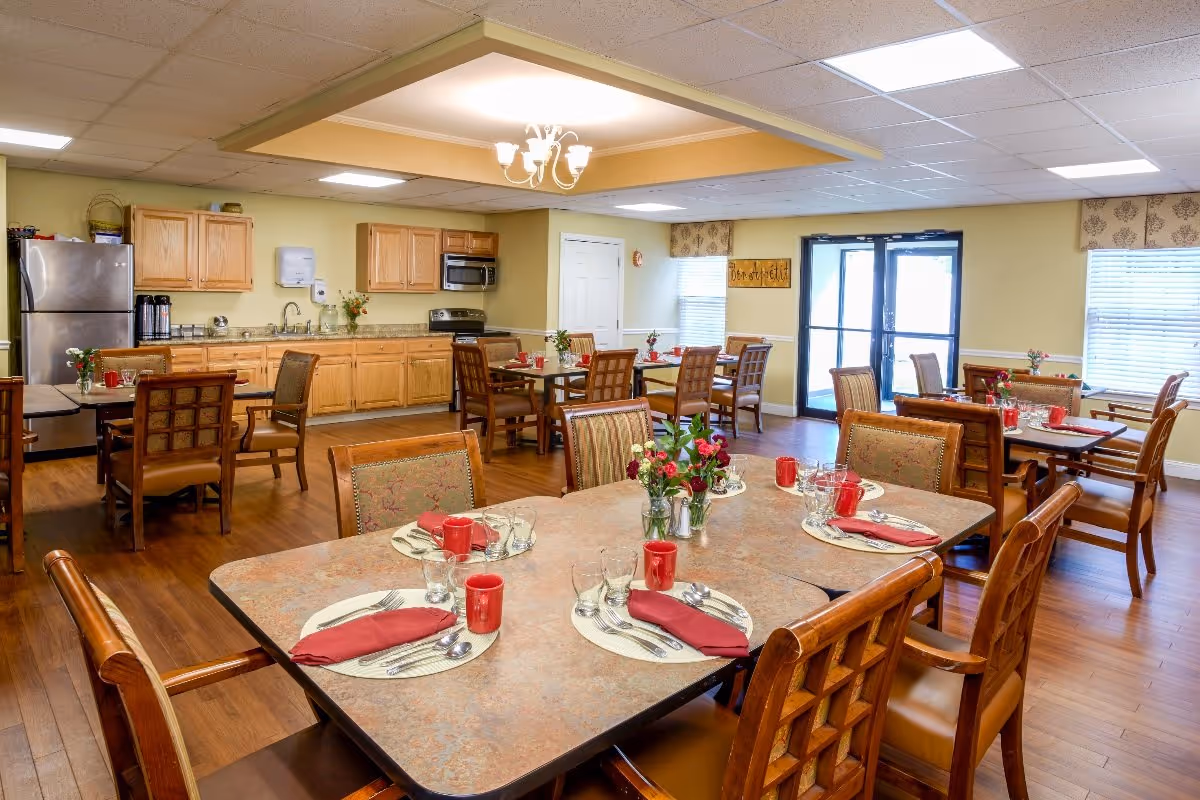 A dining room in a senior living facility with multiple tables set for meals. Each table has place settings with plates, utensils, red napkins, red mugs, and glasses. There are wooden chairs with cushioned seats around the tables. The room has a kitchen area with wooden cabinets, a stainless steel refrigerator, microwave, and stove. The floor is wooden, and there are windows with blinds and a glass door letting in natural light. A chandelier light fixture is on the ceiling.