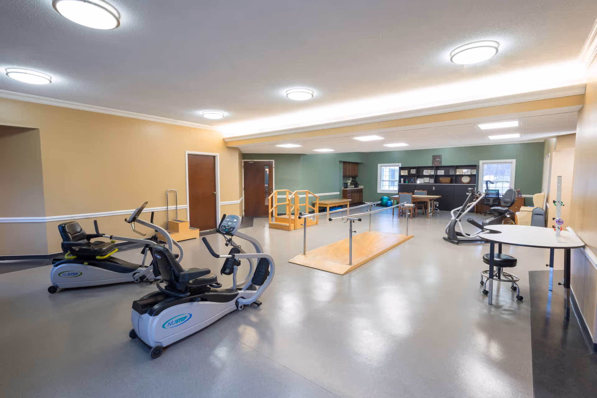 A spacious rehabilitation or physical therapy room with exercise equipment including recumbent bikes, parallel bars for walking practice, a small set of stairs, and tables with chairs. The room has beige and green walls, bright ceiling lights, and windows letting in natural light.