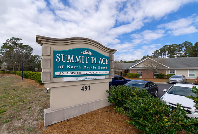 Outdoor view of the entrance sign for Summit Place of North Myrtle Beach, an assisted living community, with parked cars and residential buildings in the background under a partly cloudy sky.