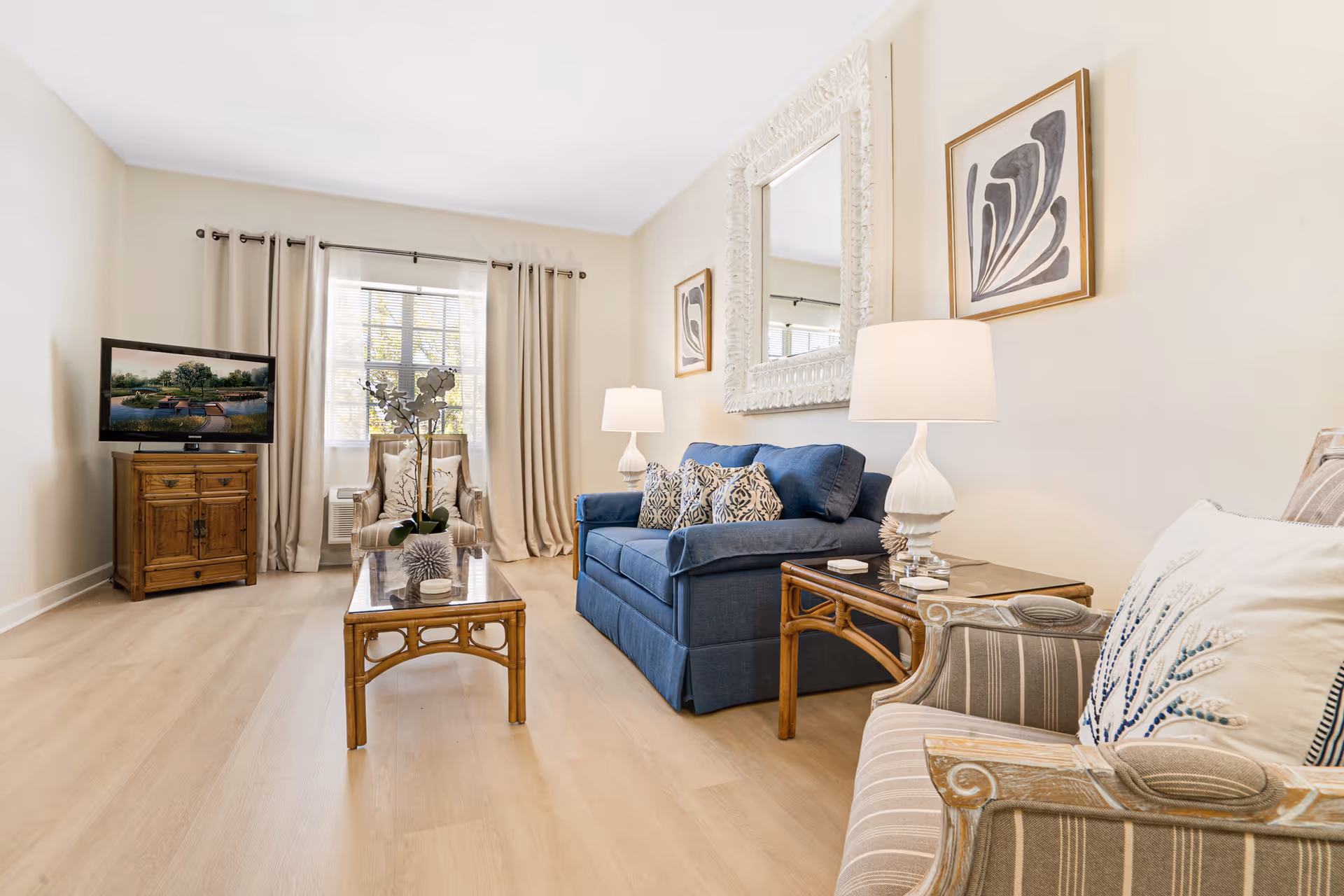 A bright living room with light wood flooring, a blue sofa with patterned cushions, two striped armchairs with decorative pillows, a wooden coffee table with a glass top and a plant centerpiece, two white table lamps on side tables, a large ornate white-framed mirror on the wall, abstract artwork, and a flat-screen TV on a wooden cabinet near a window with beige curtains.