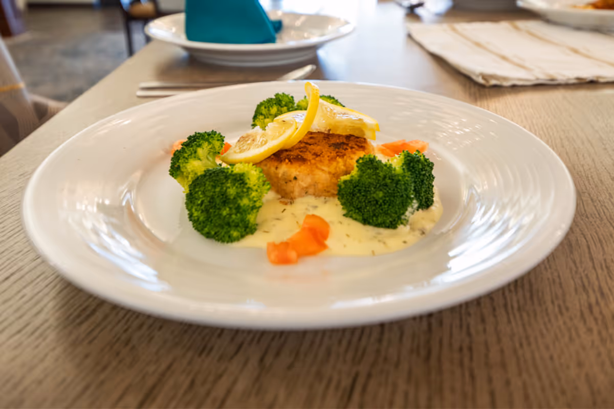 A plated meal featuring a breaded patty topped with lemon slices, surrounded by broccoli florets and small pieces of tomato, served with a creamy white sauce on a white plate on a wooden table.