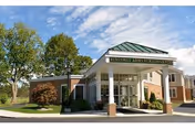 Exterior view of Kingsway Arms Nursing Center building entrance with a covered drop-off area, surrounded by trees and landscaping under a partly cloudy sky.