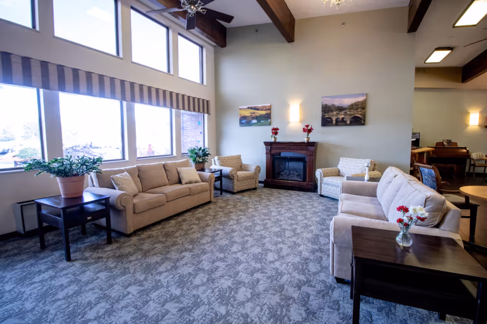 A spacious living room with large windows letting in natural light. The room features two beige sofas, two armchairs, a dark wooden fireplace with decorative flowers on top, and side tables with plants and flowers. The walls are light-colored with two landscape paintings, and the ceiling has exposed wooden beams.