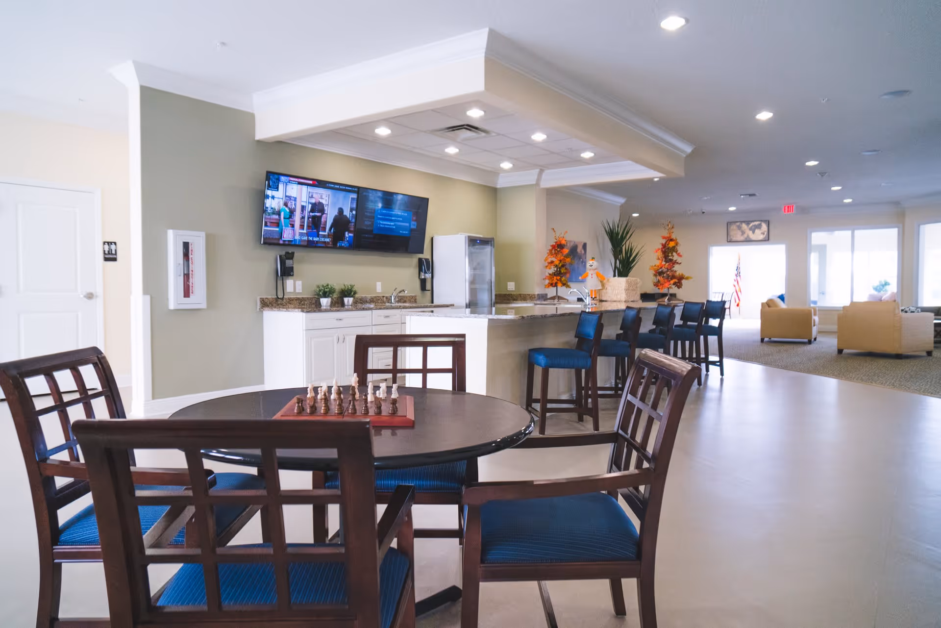 A spacious common area in a senior living facility featuring a round table with a chessboard and four chairs with blue cushions in the foreground. Behind the table is a kitchen counter with bar stools, a wall-mounted TV, and some decorative plants and autumn-themed decorations. In the background, there are beige armchairs arranged near large windows letting in natural light.