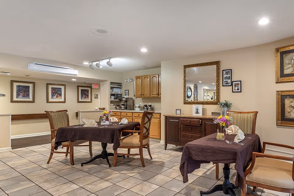 Interior view of a dining area in a senior living facility with two tables covered in dark tablecloths, each set with napkins and silverware. There are wooden chairs around the tables, a sideboard with a large mirror above it, framed pictures on the walls, and a small kitchenette area with cabinets and coffee machines in the background.