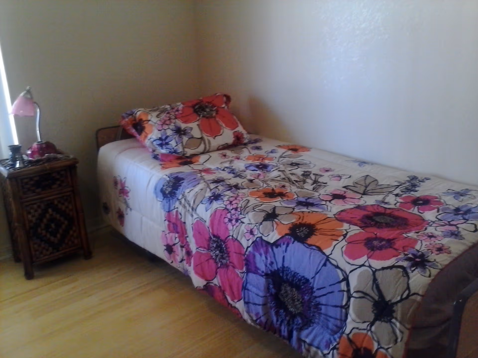 A single bed with a colorful floral bedspread and matching pillow in a simple room with light-colored walls and wooden floor. Next to the bed is a small wooden nightstand with a pink lamp and some decorative items.