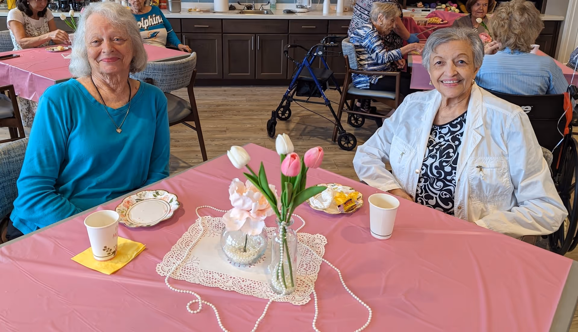Two elderly women sitting at a pink tablecloth-covered table in a dining area, smiling at the camera. The table has a vase with pink and white tulips, a small floral arrangement, and cups. Other elderly people are seated at tables in the background, with a walker visible near the back wall with cabinets.