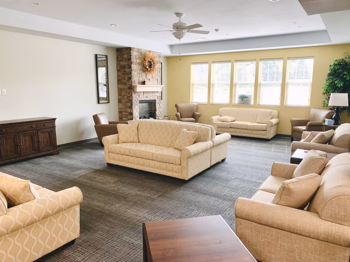 A spacious living room with multiple beige patterned sofas and armchairs arranged around a stone fireplace. The room has large windows letting in natural light, a ceiling fan, a wooden sideboard, and a potted plant with a lamp on a side table.