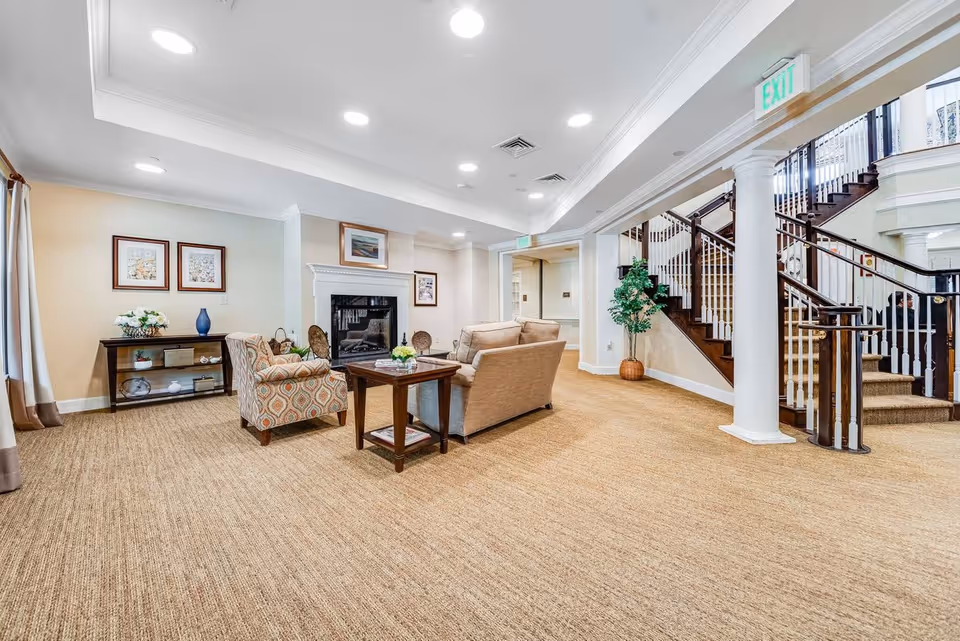 A spacious and well-lit living room area with beige carpeting, a fireplace, and comfortable seating including a sofa and an armchair. There is a wooden coffee table in the center with a small plant on it. The room features framed artwork on the walls, a console table with decorative items, and a staircase with dark wood railings and white balusters leading to an upper floor. A potted plant is placed near the staircase, and an exit sign is visible above a doorway.