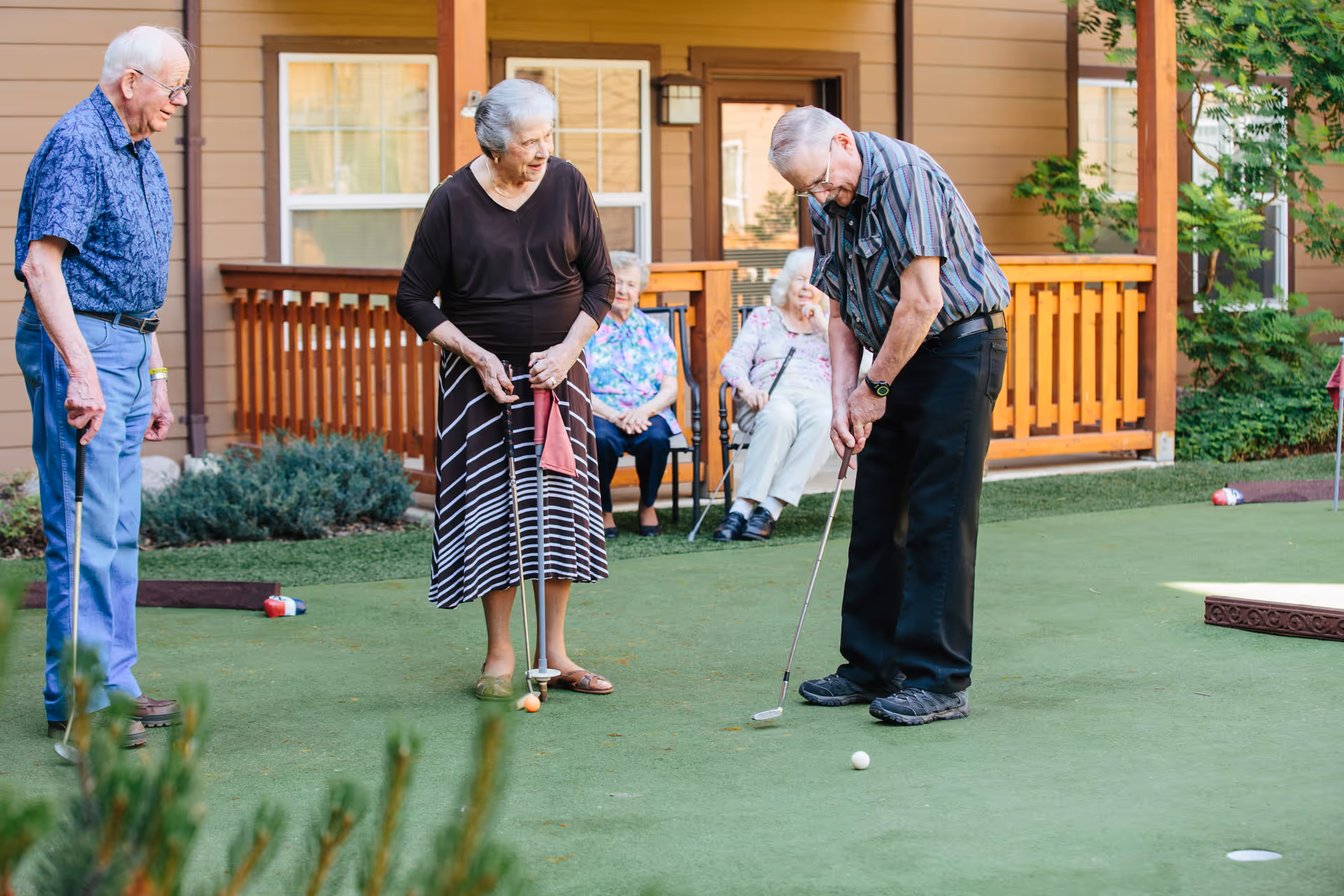 Three elderly people playing mini golf on a green putting surface outside a building with wooden railings. Two elderly women are sitting on chairs in the background watching the game.