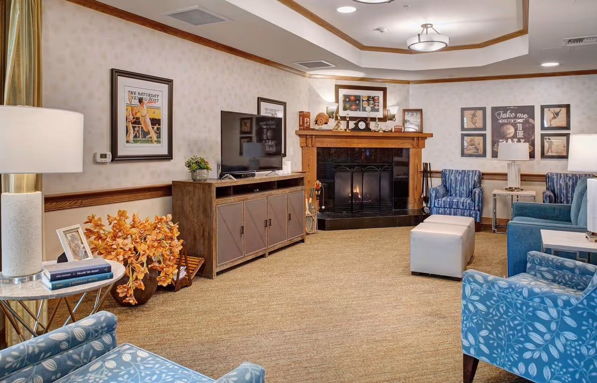 A cozy living room with a fireplace, a flat-screen TV on a wooden cabinet, blue patterned armchairs, a blue sofa, two white ottomans, framed artwork on the walls, a side table with a lamp and books, and a vase with orange flowers on the floor.